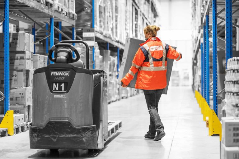 Une femme qui porte un carton à côté d'un chariot élévateur dans une allée d'un entrepôt de logistique